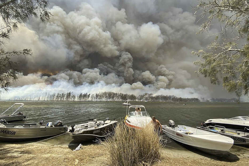 Boote werden an Land gezogen, während im Hintergrund Rauchwolken über dem Ufer hinter dem Canjola See aufsteigen.