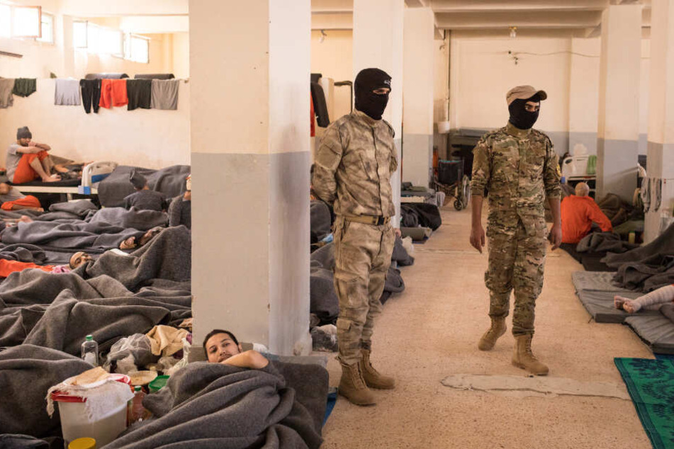 Zwei Männer bewachen zahlreiche Gefangene in orangenen Overalls, die in einem Raum in Hassake (Syrien) sitzen und liegen.