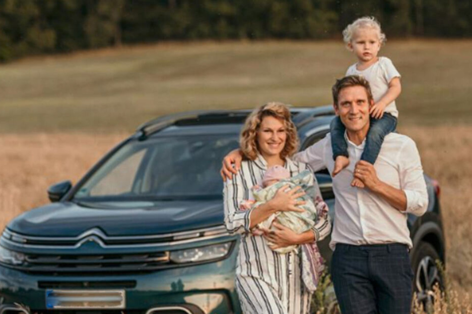Janni und Peer stehen mit ihren Kindern vor ihrem Auto auf einem Kornfeld.