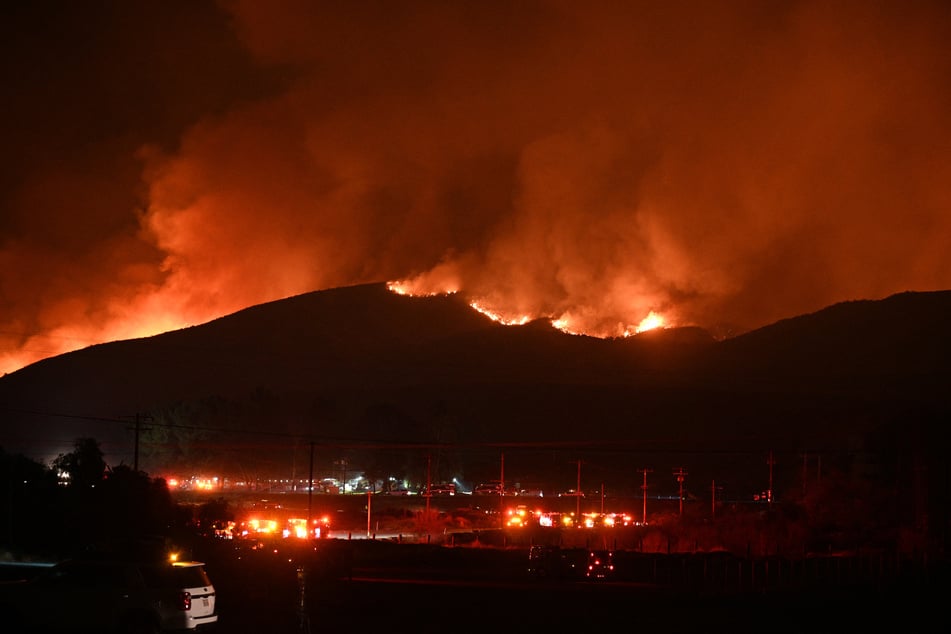 Fire trucks and public security vehicles are seen in the foreground as red glowing flames and smoke rise from the Hughes Fire in Castaic in northwest Los Angeles County, California, on January 22, 2025.