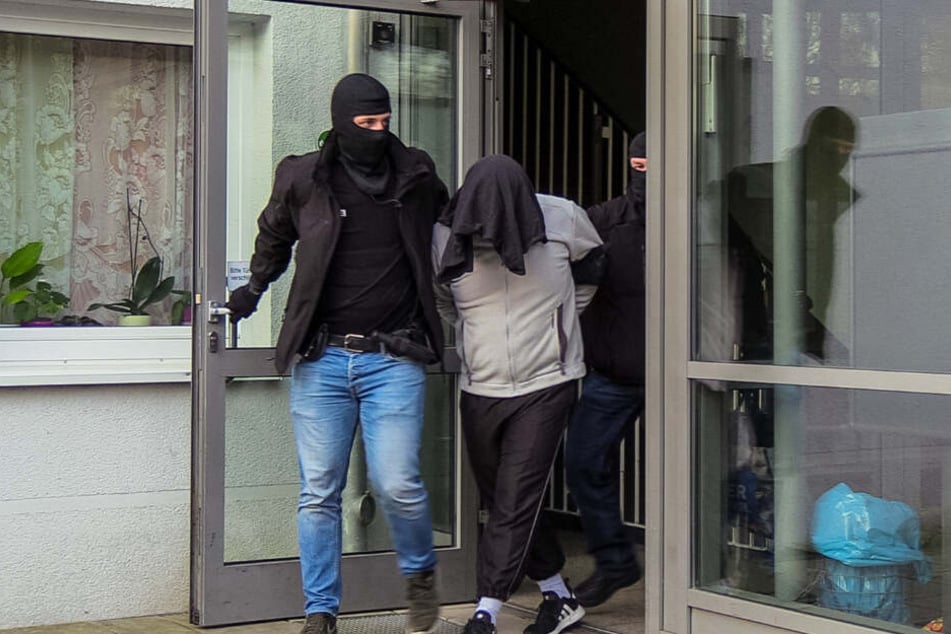 Der Tatverdächtige wird von zwei vermummten Beamten aus dem Haus geführt.