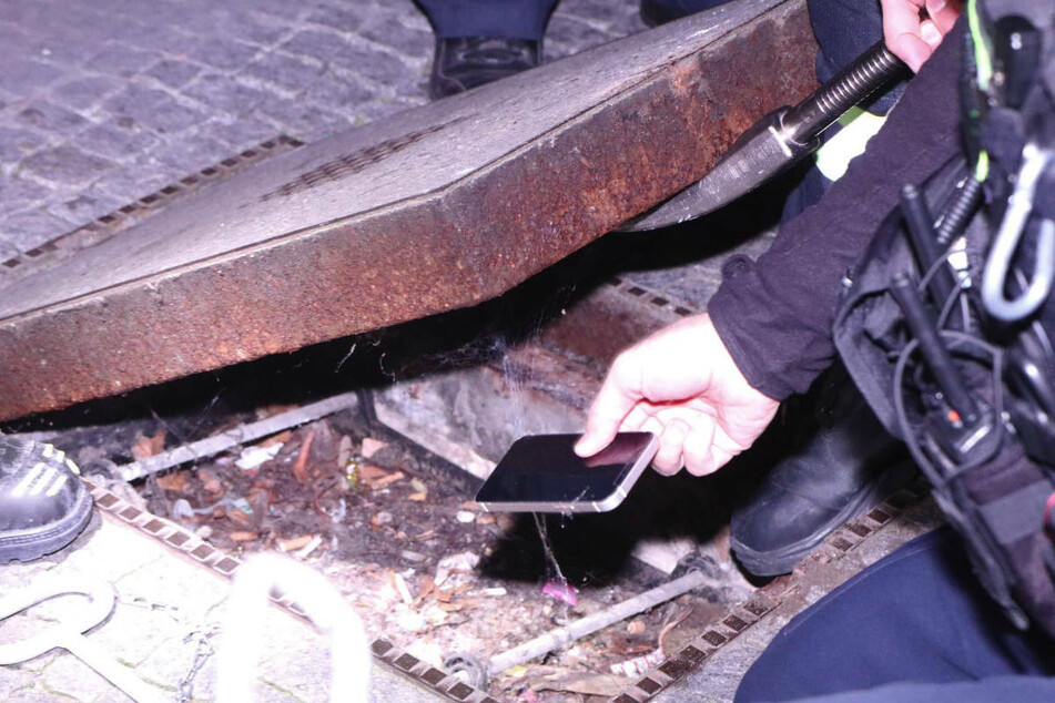 Ein Polizist hebt das Smartphone auf, das tatsächlich unter dem schweren Deckel zum Vorschein gekommen ist.
