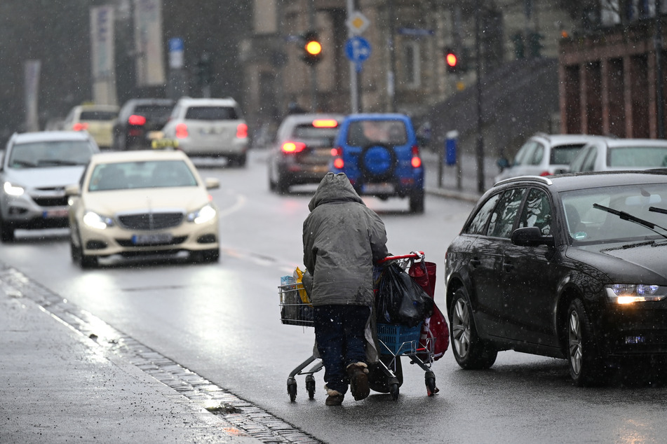 Autos weichen einer obdachlosen Frau aus, die ihr Hab und Gut in einem Einkaufswagen am Rande einer Fahrbahn einer Straße schiebt.