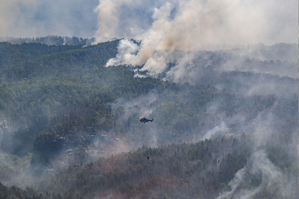 Ein Lastenhubschrauber aus Österreich fliegt mit einem Löschwasser-Außenlastbehälter, um einen Waldbrand im Nationalpark Sächsische Schweiz zu löschen.