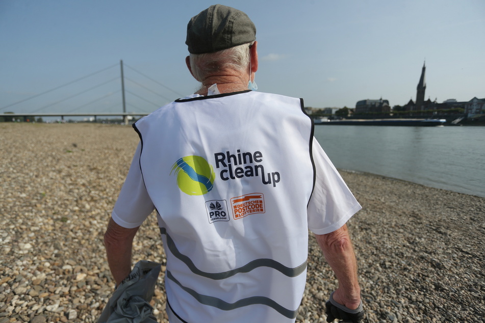 in Mann, der den Schriftzug "Rhine Clean Up" auf seinem Rücken trägt, geht am Rheinufer entlang.