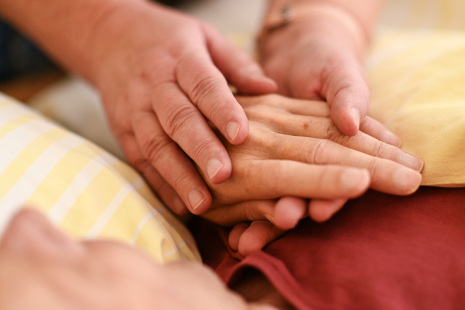 Eine Hospizmitarbeiterin hält die Hand eines todkranken Menschen, der in einem Hospiz im Bett liegt. (Symbolfoto)