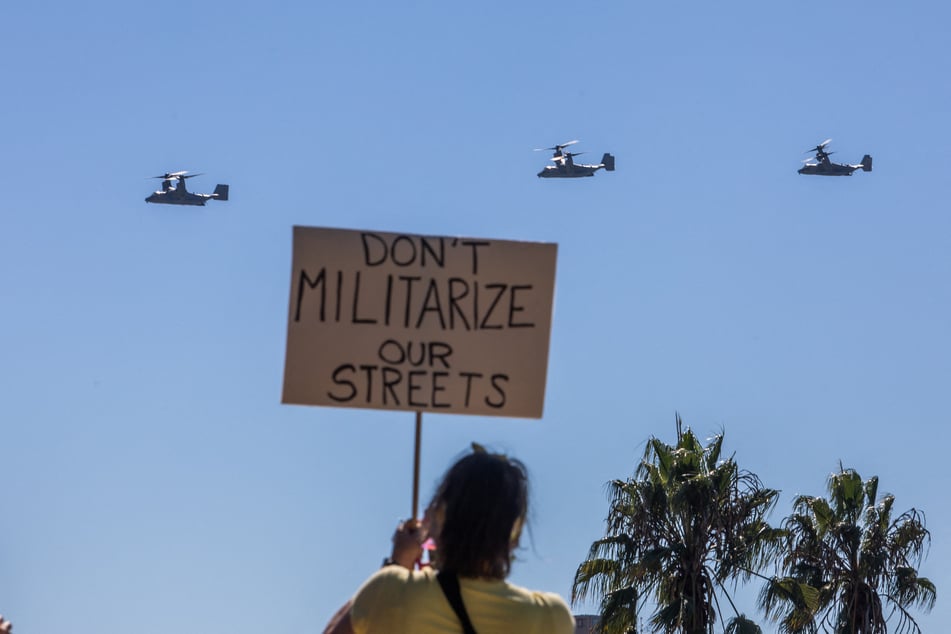 Protesters hold signs at the main entrance of the Marine Corps Base Camp Pendleton as US Marine helicopters fly overhead on October 18, 2025.