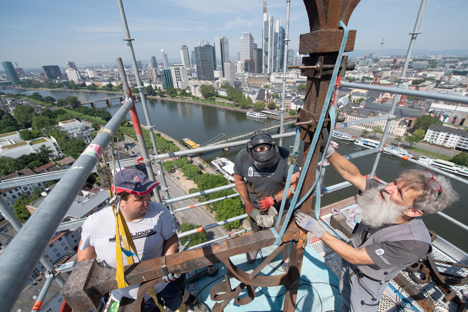 Vor der Hochhauskulisse von Frankfurt arbeiten Maurer Jonas Kirchner, Metallbauer David Pößel und Steinmetz Reinhart Vierkant (v.l.n.r.) an der Demontage des Kreuzes auf dem Turm der Dreikönigskirche am Mainufer.
