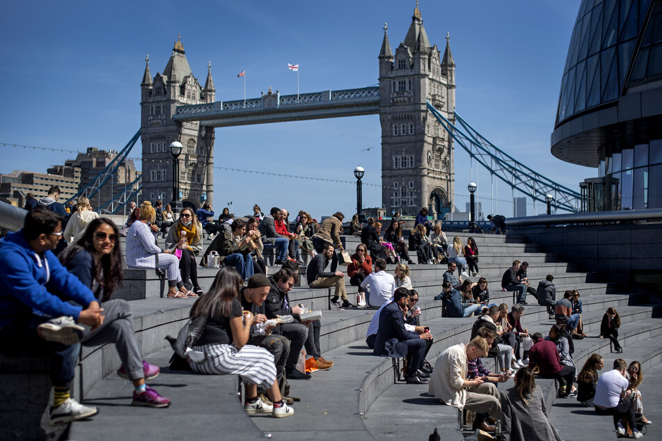 Zahlreiche Menschen sitzen bei gutem Wetter in der Nähe der Tower Bridge in London auf Treppenstufen. Großbritannien kommt im Kampf gegen das Coronavirus gut voran, zahlreiche Menschen sind bereits geimpft.