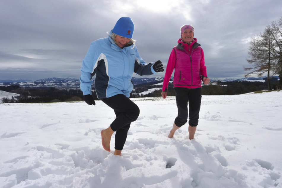 Ida-Anna Braun (l.,69), Vorsitzende des Kneipp-Vereins Kempten, und Monika Weitnauer, Schriftführerin und Nordic-Walking-Leiterin des Vereins, gehen vor dem Panorama der Alpen barfuß durch den Schnee.