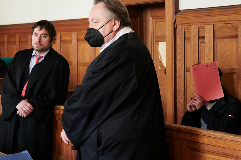 Der Angeklagte Leonardo D. hält zu Prozessbeginn im Saal des Amtsgerichts Berlin-Tiergarten eine Pappe vor sein Gesicht, seine Verteidiger Bernd Kubacki (M) und Georg Weber stehen vor ihm.