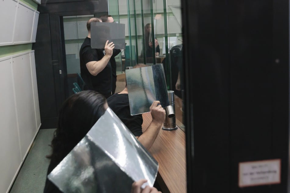 Die vier Angeklagten verdecken ihre Gesichter hinter silberfarbenen Ordnern.