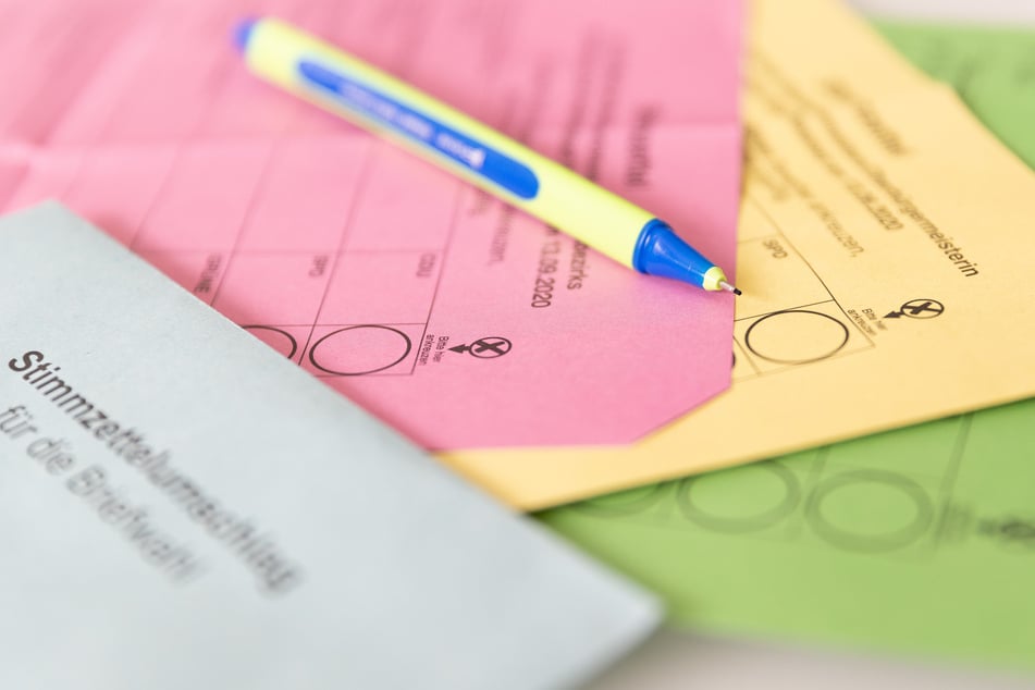 Verschiedene Stimmzettel und ein Stimmzettelumschlag für die Briefwahl liegen auf einem Tisch zusammen mit einem Stift.