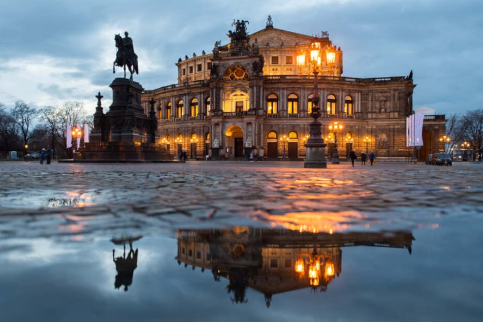 Die Semperoper und das König-Johann-Denkmal spiegeln sich am Abend in einer Pfütze auf dem Theaterplatz.