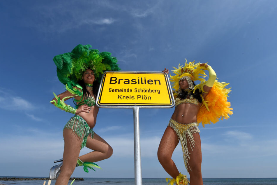 Die brasilianische Tänzerinnen Luciana (links) und Simone stehen neben dem Ortsschild von Brasilien am Ostseestrand. Der Strandabschnitt in der Gemeinde Schönberg ist einer von vielen Ortsnamen in Schleswig-Holstein, die nach weiter Welt klingen.