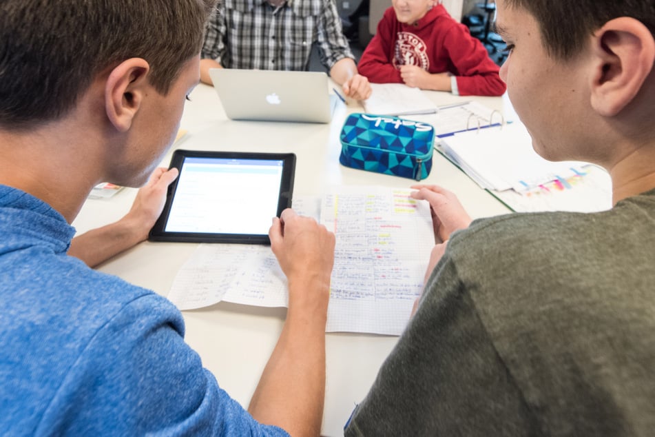 Schüler und ein Lehrer sitzen an einer Gesamtschule vor ihren Tablets und einem Laptop.