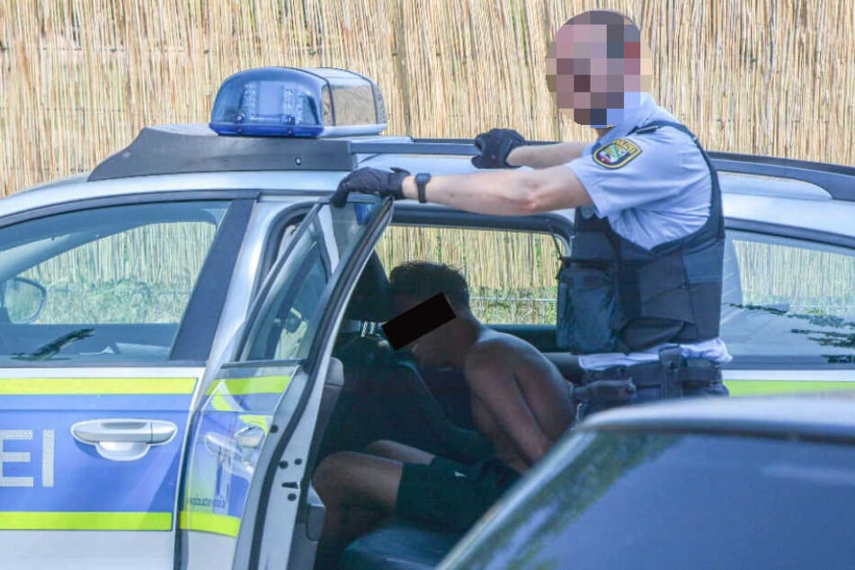 Einer der Täter sitzt nach dem Angriff in Handschellen in einem Polizeiauto und wird von einem Beamten bewacht.