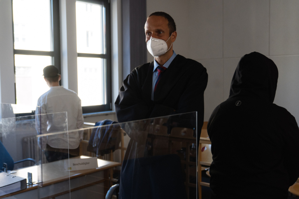 Zwei von drei Angeklagten warten im Gerichtssaal auf den Prozessbeginn. In der Mitte steht der Verteidiger Christian Reimesch.