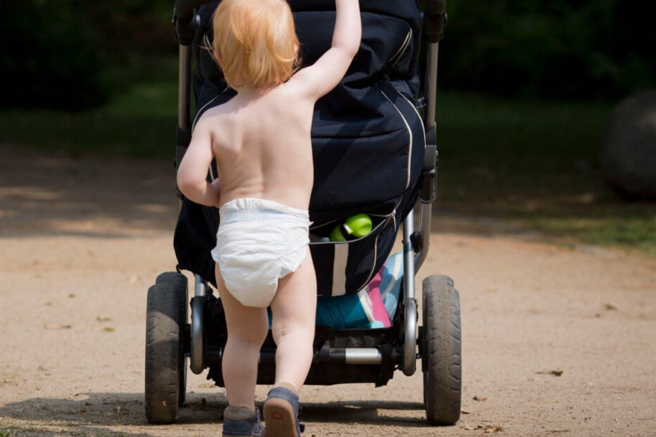 Ein nur mit einer Windel bekleidetes Kind schiebt einen Kinderwagen.