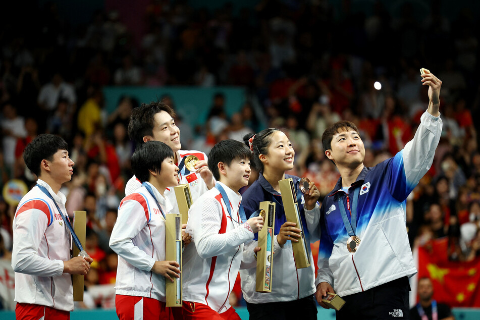 Bronze medalist Jonghoon Lim of South Korea takes selfie with Yubin Shin of South Korea and gold medalists Chuqin Wang and Yingsha Sun of China with silver medalists Jong Sik Ri and Kum Yong Kim of North Korea on the podium.