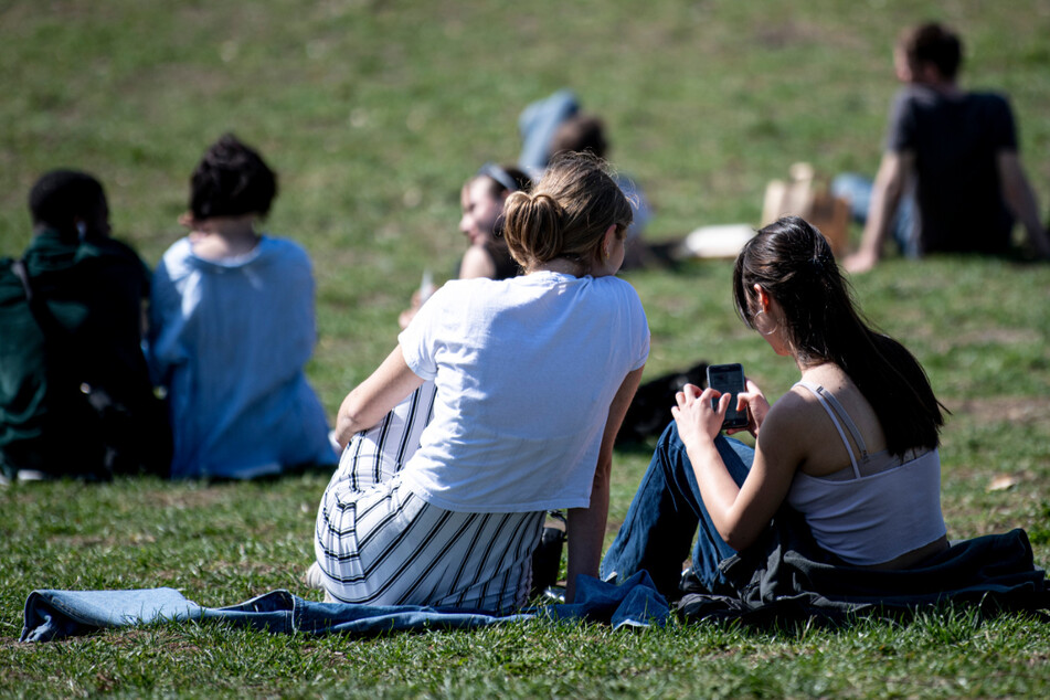 Zwei Frauen sitzen bei warmen Temperaturen im Mauerpark auf einer Wiese und blicken auf ein Smartphone.