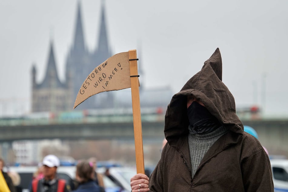 Ein als Sensenmann verkleideter Teilnehmer einer Demonstration gegen Corona-Schutzmaßnahmen trägt ein Sense aus Pappe mit der Aufschrift "gestorben wird immer".