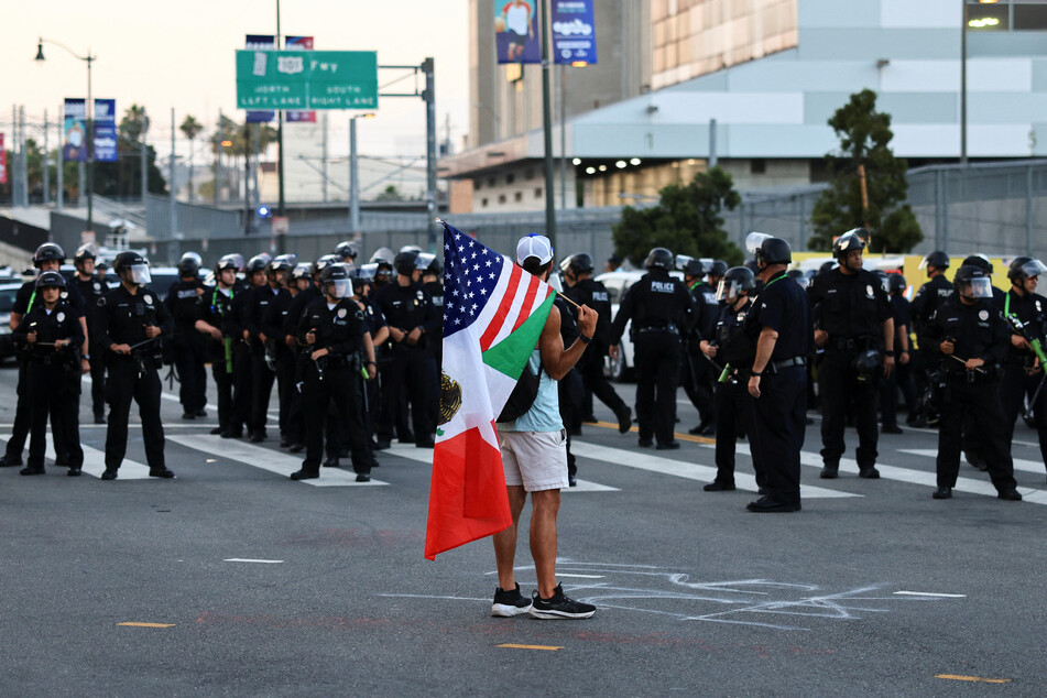 A person holds a mixed Mexico-US flag as law enforcement officers stand at the ready during a protest against federal immigration raids in Los Angeles, California, on June 12, 2025.