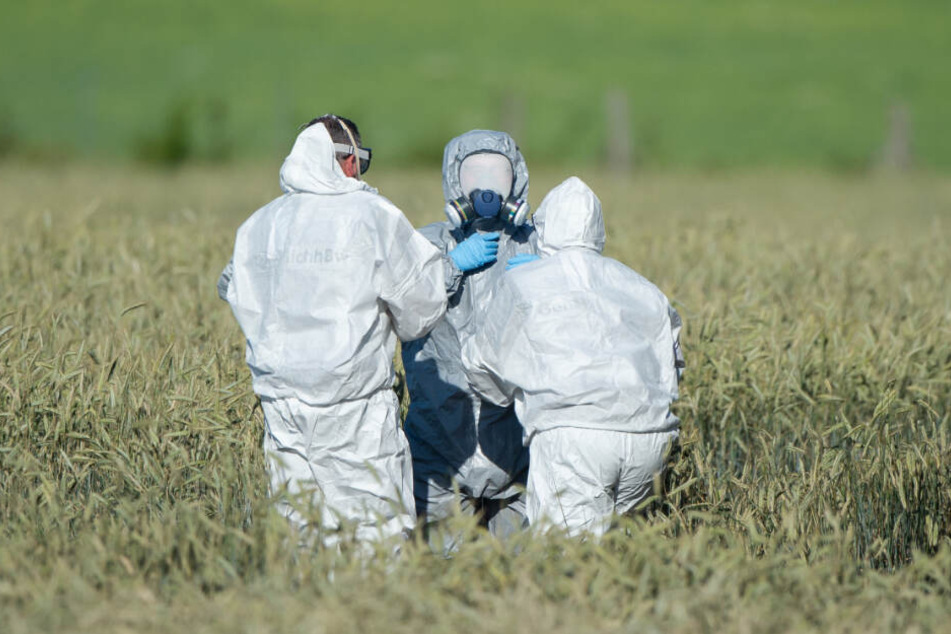 Mitarbeiter der Flugsicherheit der Bundeswehr helfen einem Mitarbeiter aus dem Schutzanzug, der auf einem Feld Wrackteile untersucht hat.
