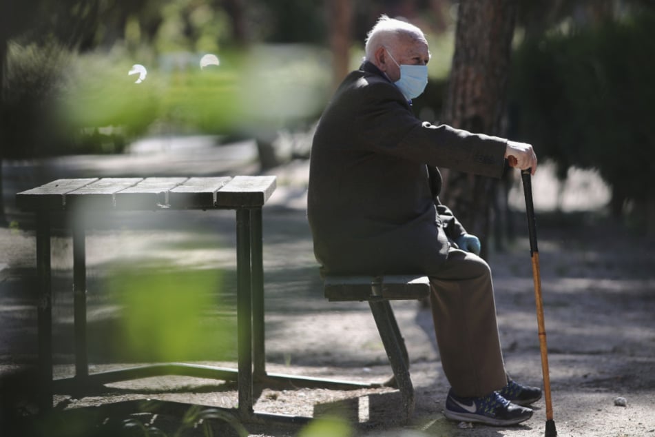 Ein älterer Mann mit einer medizinischen Mundschutzmaske sitzt auf einer Bank in einem Park. (Symbolbild)