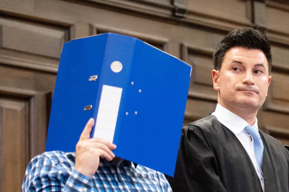 Der Angeklagte verbirgt sein Gesicht zu Prozessbeginn hinter einem Aktenordner. Daneben steht sein Verteidiger Haydar Güler.