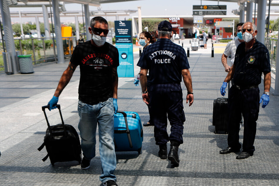 Ein Mann mit Mundschutz und Reisekoffern geht am Flughafen Larnaka an Polizisten vorbei.