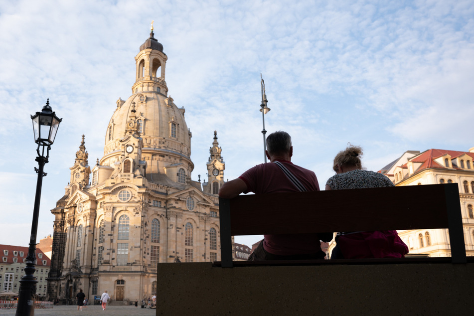 Passanten sitzen auf dem Neumarkt vor der Frauenkirche auf einer Bank.