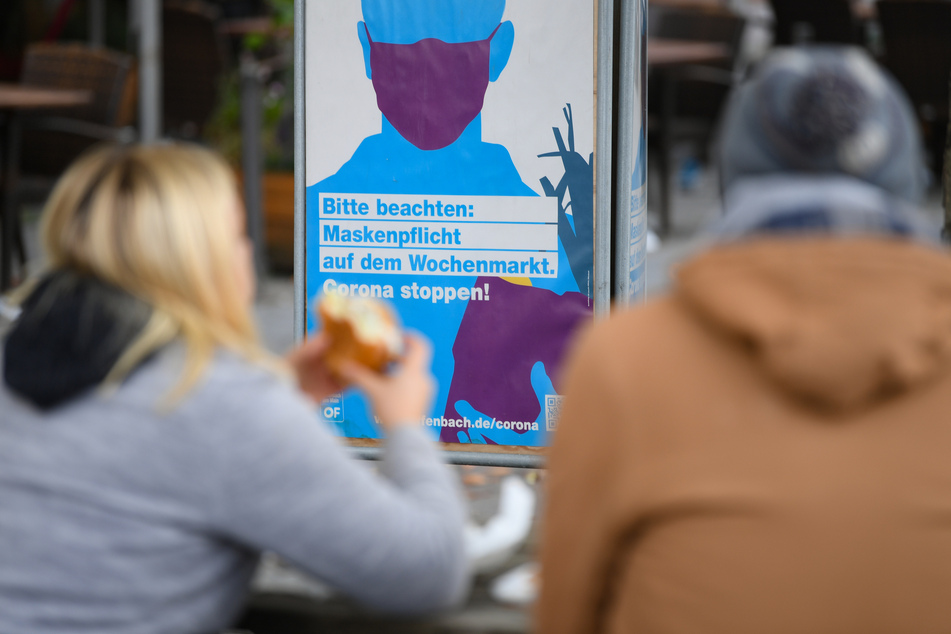 Ein Paar sitzt auf dem Marktplatz von Offenbach vor einem Plakat mit der Aufschrift "Bitte beachten: Maskenpflicht auf dem Wochenmarkt. Corona stoppen!".