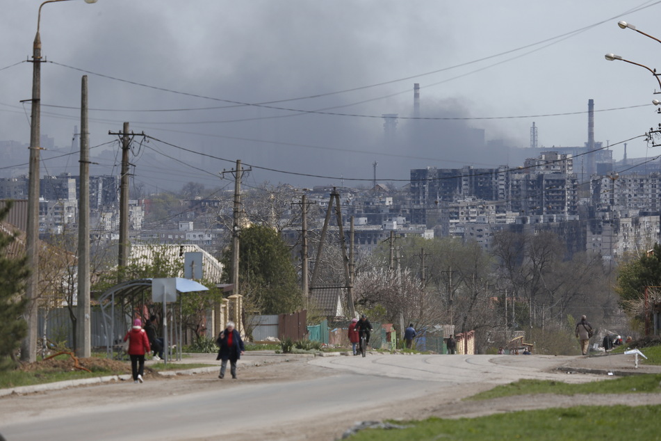 Anwohner gehen über die Straße, während im Hintergrund Rauch aus dem Azovstal-Werk in der Hafenstadt Mariupol aufsteigt. Russische Truppen haben die Angriffe auf das Stahlwerk wieder aufgenommen.