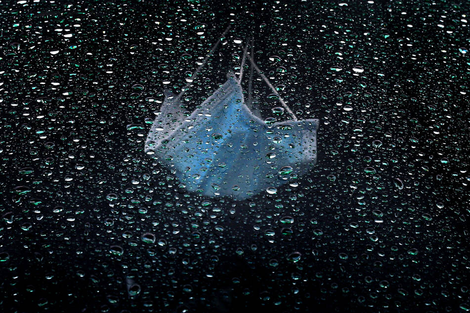 Ein medizinischer Mundnasenschutz hängt am Rückspiegel eines im Regen parkenden Autos.