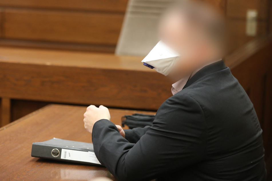 Ein wegen Volksverhetzung und öffentlichem Aufruf zu Straftaten angeklagter Polizeibeamter sitzt mit Mund- Nasenschutz im Gerichtssaal.