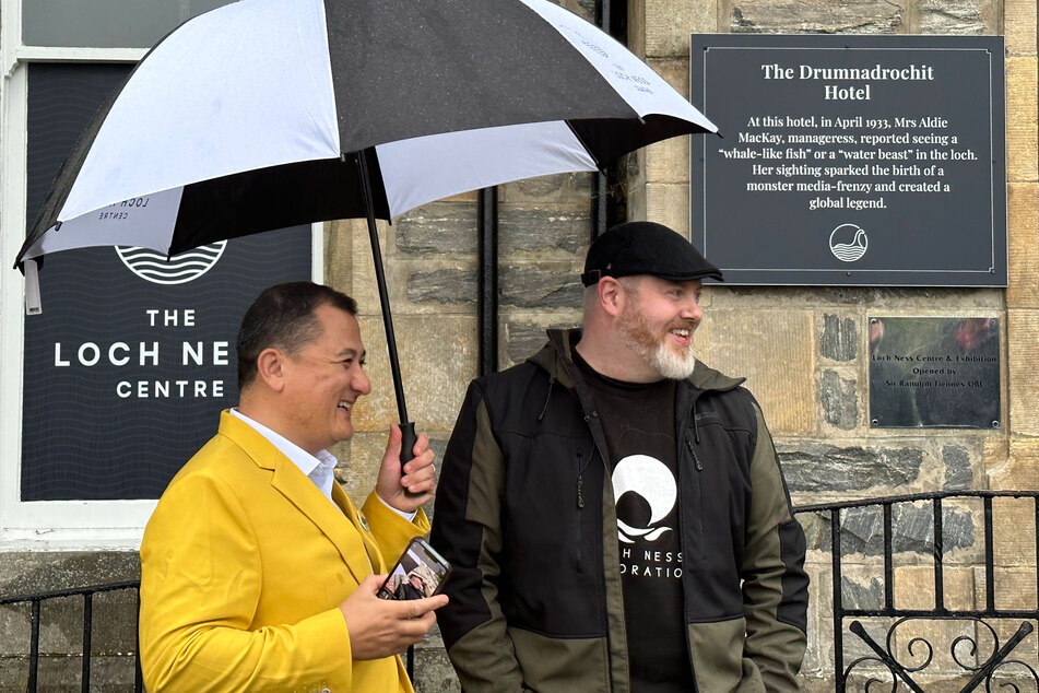 Paul Nixon (l.) vom Loch Ness Centre und Projektleiter Alan McKenna stehen vor der Touristenattraktion "Loch Ness Centre" neben einer Plakette, die an eine berühmte Nessie-Sichtung im Jahr 1933 erinnert.