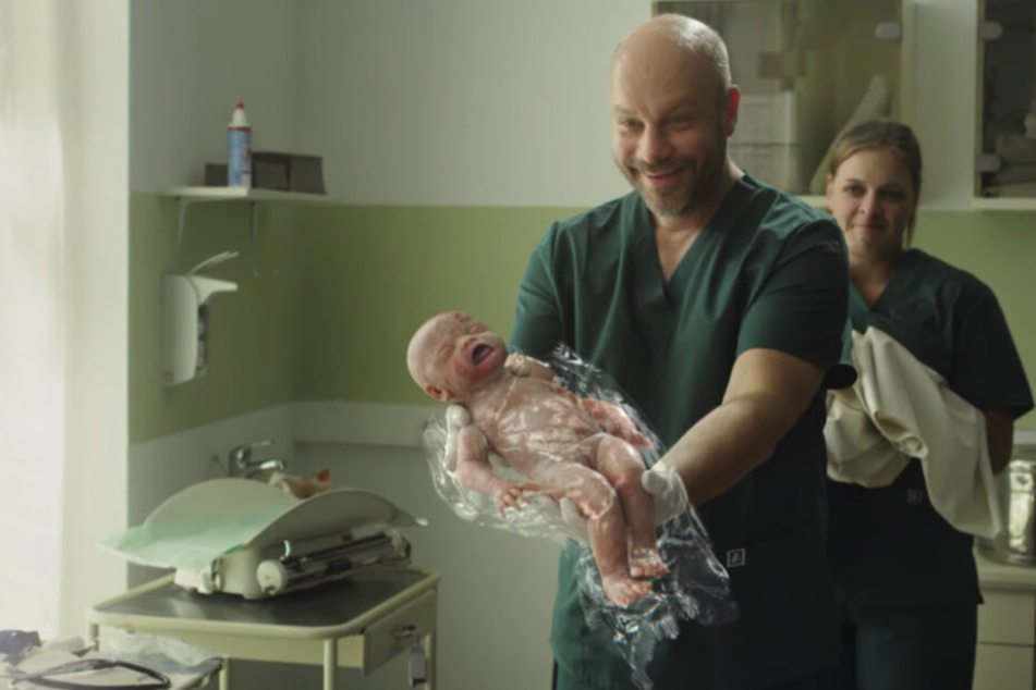 Ein Arzt hält das Kind nach der Geburt in Plastik eingehüllt vor die Eltern.