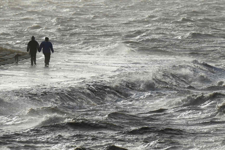 Wellen der aufgepeitschten Nordsee schlagen gegen einen Deich. Zwei Spaziergänger stört das nicht. (Archivbild)