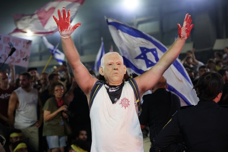 A protester, wearing a mask depicting Israel's Prime Minister Benjamin Netanyahu, raises his hands wearing gloves with red paint during an anti-government rally.