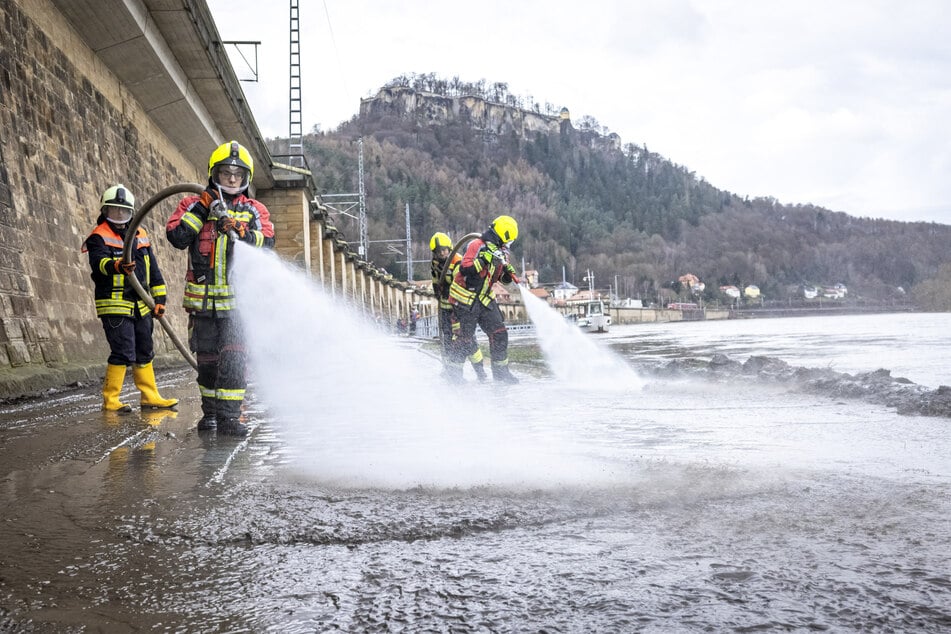 In Königstein reinigen Feuerwehrleute eine Straße entlang der Elbe vom angespülten Schmutz.