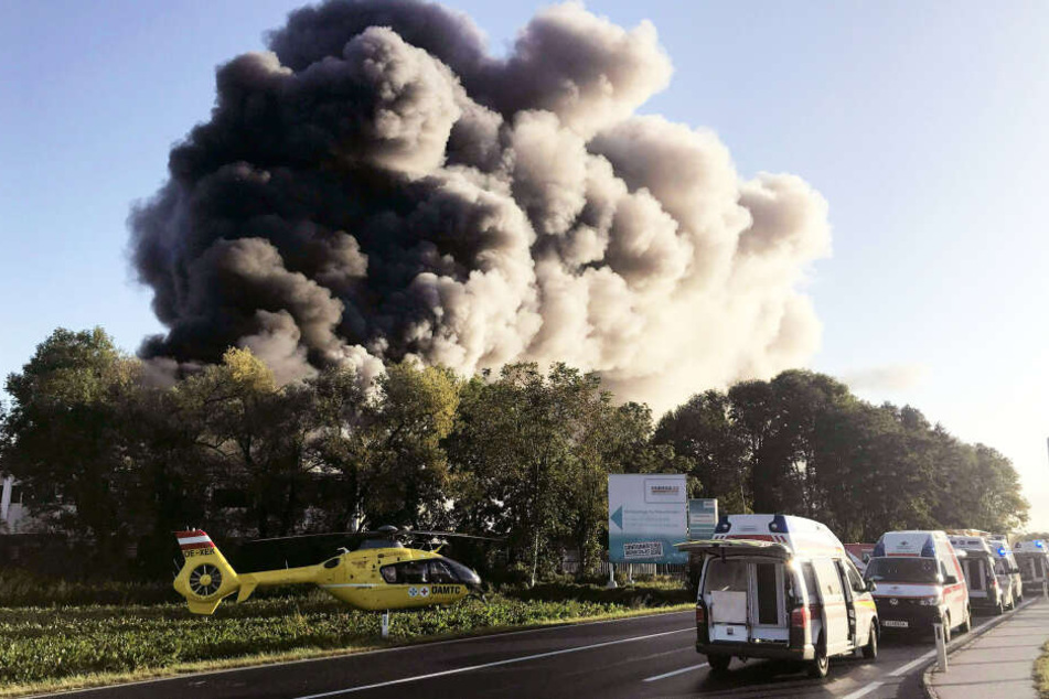 Eine Rauchwolke steigt hinter Bäumen in den Himmel empor, auf der Straße davor stehen Rettungswagen und ein Rettungshubschrauber.