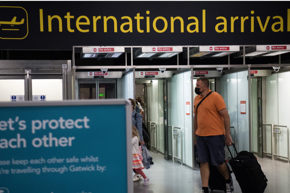 Passagiere kommen am Flughafen Gatwick in West Sussex an, an dem ein Schild mit der Aufschrift "Let's protect each other" (Lasst uns aufeinander aufpassen) steht.