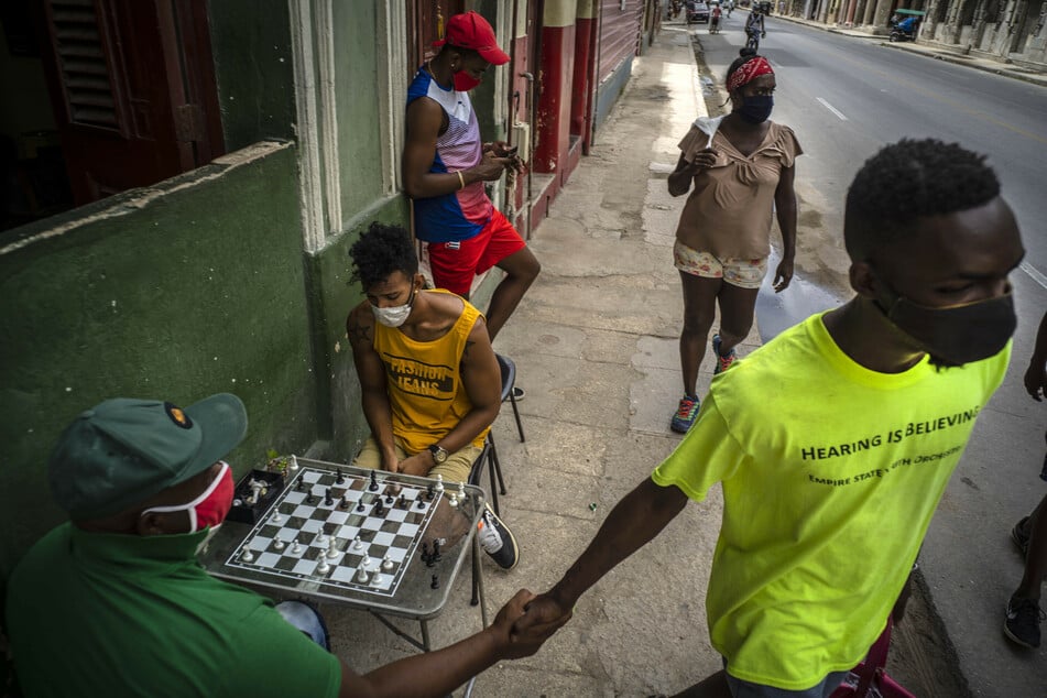 Havanna: Menschen mit Mundschutz gehen auf einer Straße und schütteln andere, die auf einem Bürgersteig Schach spielen, die Hand.