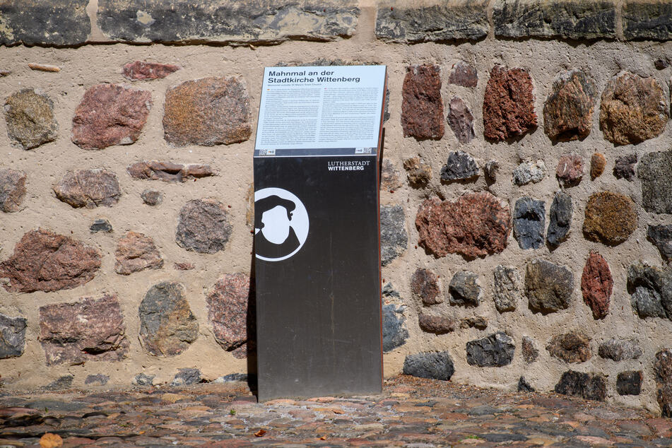 Eine Infotafel mit der Aufschrift "Mahnmal an der Stadtkirche Wittenberg" ist am Fuße der Stadtkirche von Wittenberg zu sehen. Der Text informiert auf Deutsch und in Englisch über das antijüdischen Schmähplastik.