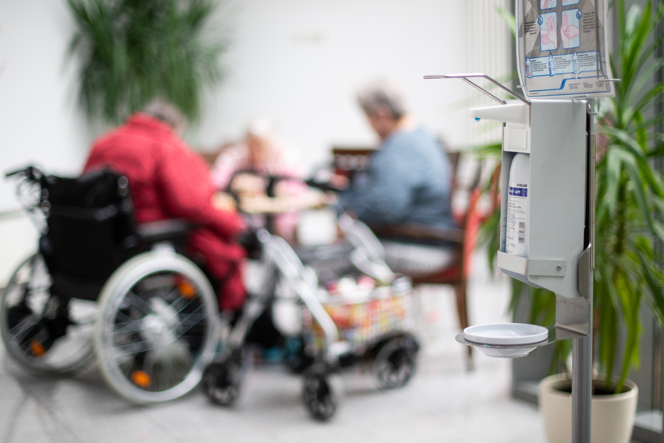 Ein Desinfektionsmittelspender hängt in einem Altenzentrum, während im Hintergrund Seniorinnen spielen.