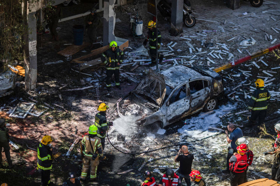 Im israelischen Ramat Gan arbeiten Feuerwehrleute an einem Autowrack vor einem Wohnhaus, das im Mai 2021 von einer Rakete der islamistischen Hamas getroffen wurde. Zwei Monate nach den Raketenangriffen auf Israel kommen 60 israelische Kinder und Jugendliche zur Erholung nach Berlin.