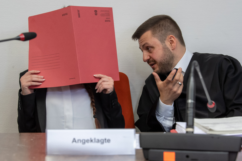 Die Angeklagte hält auf ihrem Sitz im Gerichtssaals einen roten Aktendeckel vor ihr Gesicht. Rechts spricht ihr Anwalt Ali Aydin mit ihr. (Archiv)