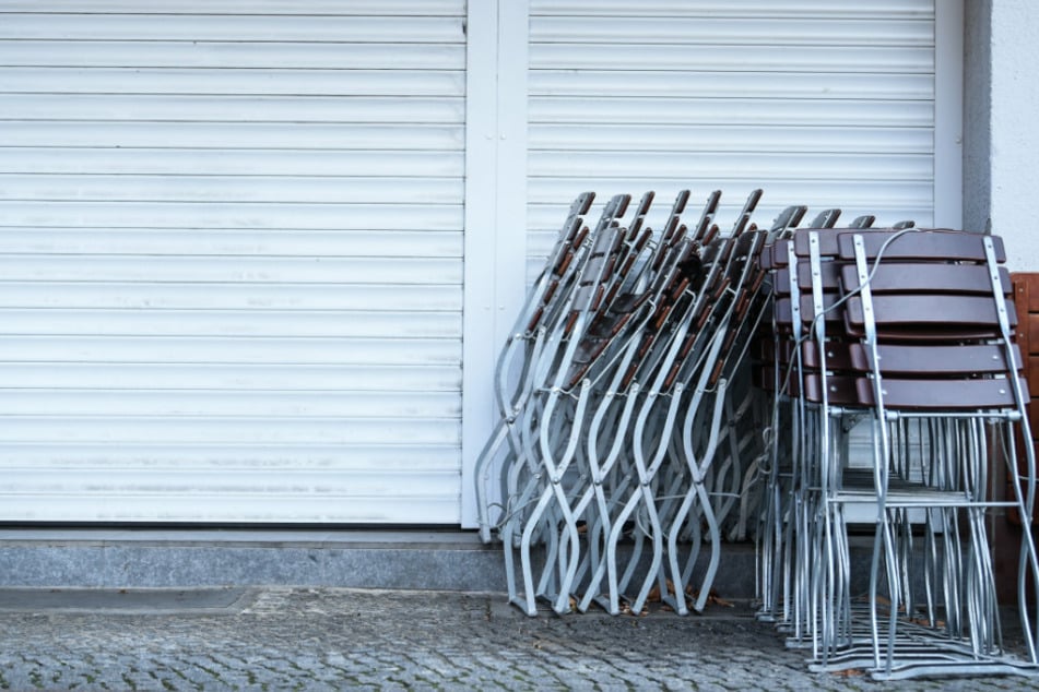 Vor einem Geschäft stehen zusammen gestapelte Stühle und die Rollläden sind heruntergelassen.