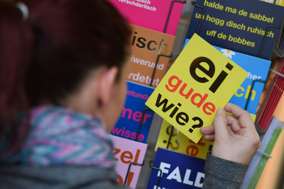 Eine Frau hält vor dem Hessen-Shop in der Frankfurter Kleinmarkthalle eine Postkarte mit dem klassischen hessischen Satz "ei gude wie?" (Symbolbild).